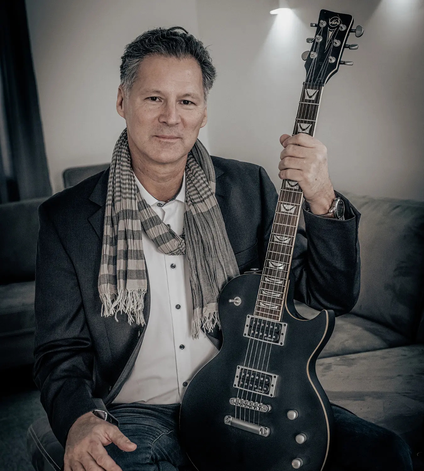 Auf diesem Bild sehen Sie einen Mann, der auf einem Sofa sitzend stolz seine elektrische Gitarre in der Hand hält