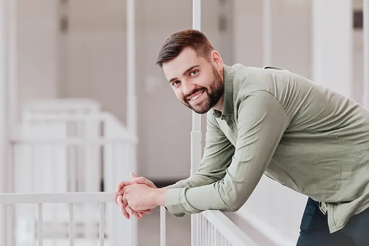 Auf diesem Bild sehen Sie einen jungen Mann mit Hemd und Jeans bekleidet, der auf einem Geländer in einem Firmengebäude lehnt