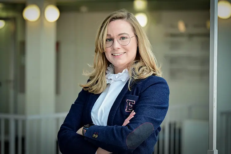 Auf diesem Bild sehen Sie eine junge Frau, die selbstbewusst ihre Arme verschränkt und in die Kamera lächelt