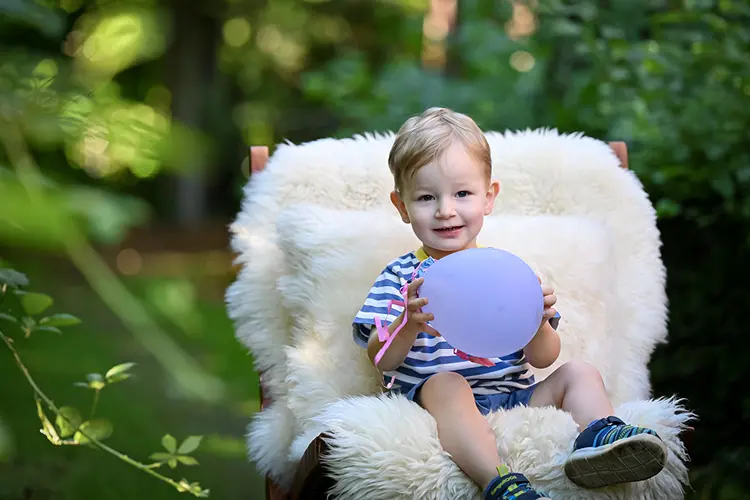 Auf diesem Bild sehen Sie einen kleinen Jungen auf einem Stuhl sitzend, der in die Kamera lächelt und dabei Luftballons in der Hand hält