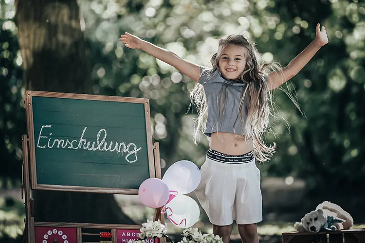 Auf diesem Bild sehen Sie ein junges Mädchen am Tag ihrer Einschulung, was neben einer Schultafel vor Freude in die Luft springt