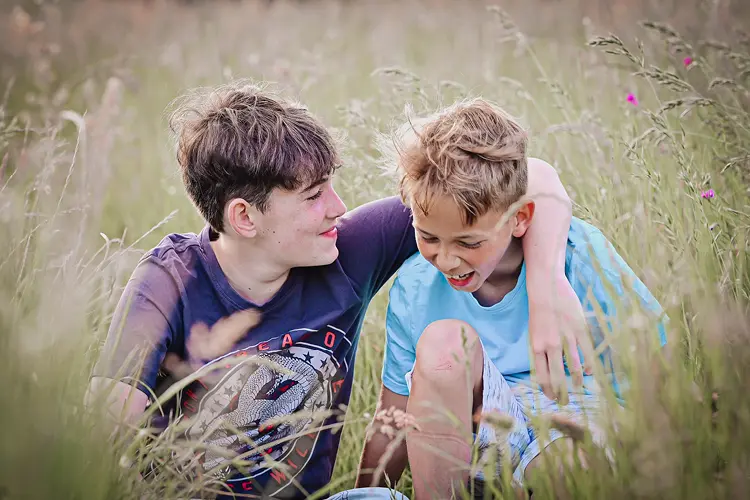 Auf diesem Bild sehen Sie zwei Kinder, die sich in einem Blumenfeld sitzend lachend umarmen