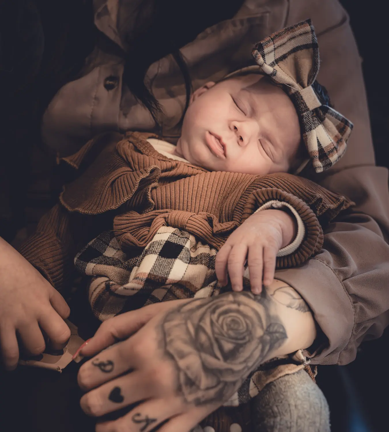Auf diesem Bild sehen Sie ein kleines Baby mit einer Schleife auf dem Kopf, was in den Armen seiner tattoowierten Mama schläft