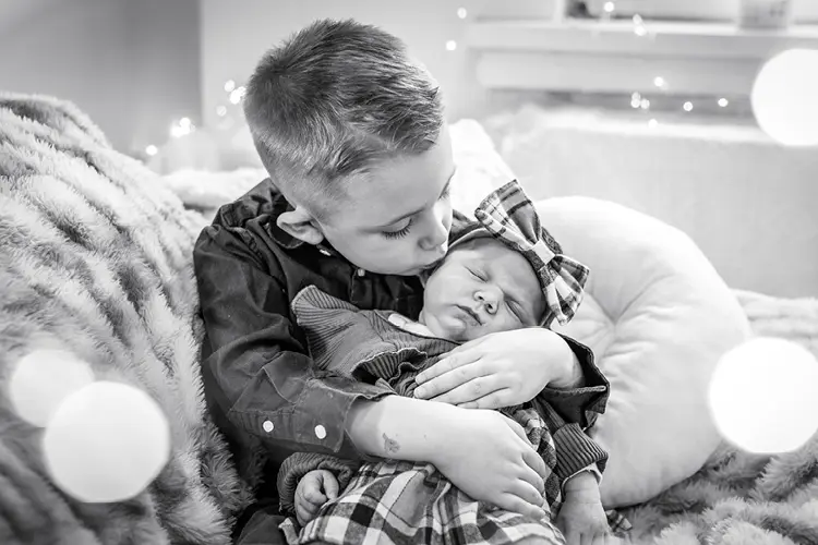 Auf diesem Bild sehen Sie ein kleines Baby mit einer Schleife auf dem Kopf, was von seinem kleinen Bruder schlafend einen Kuss auf die Wange bekommt