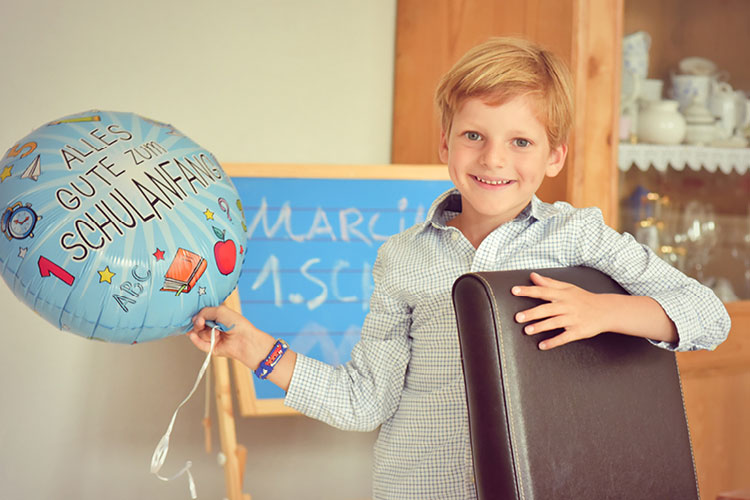 Auf diesem Bild sehen Sie einen jungen an seiner Einschulung mit einem Luftballon in der Hand neben einer Schultafel