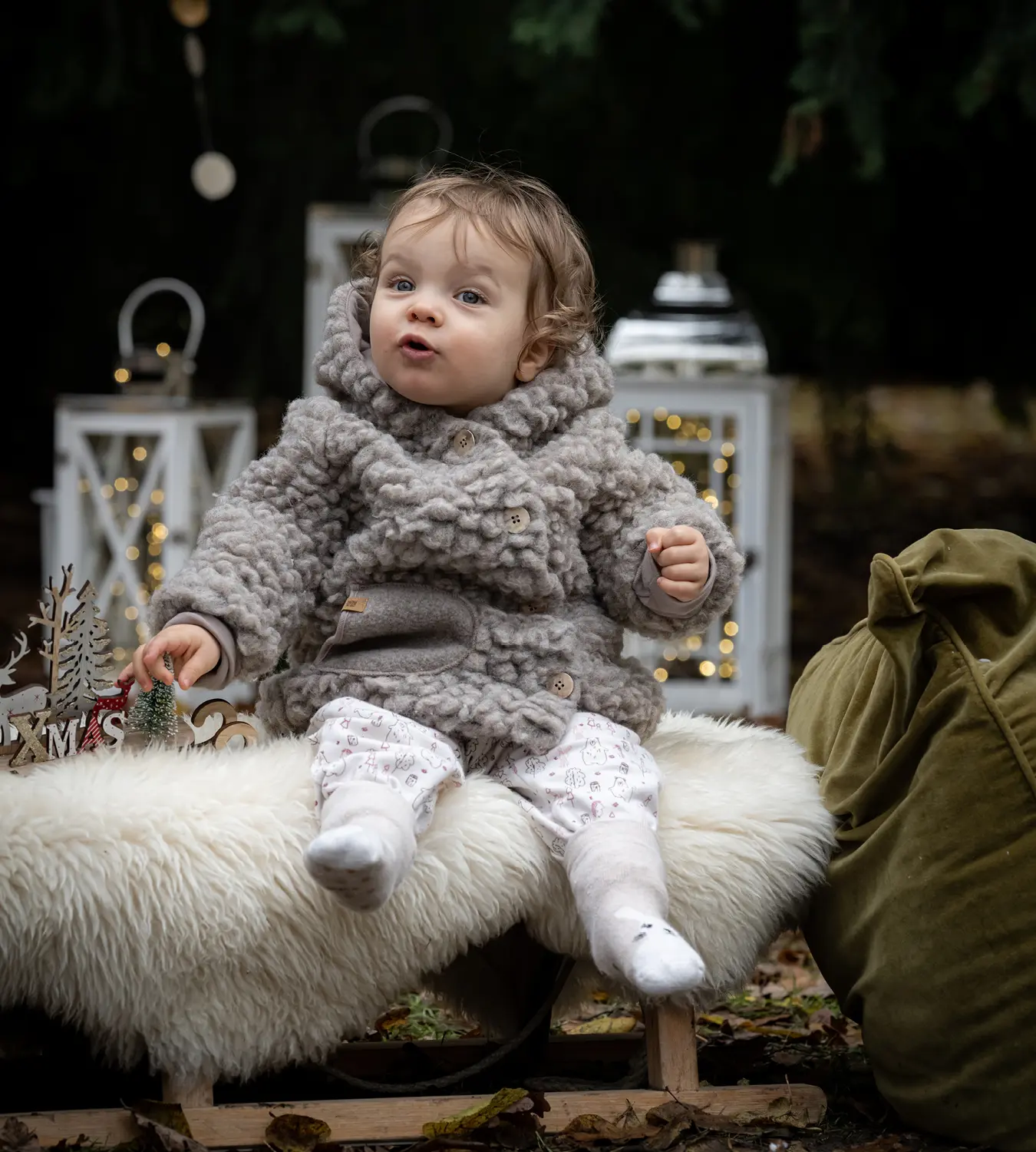 Auf diesem Bild sehen Sie ein Baby, was in winterlicher Atmosphäre vor Laternen sitzend dick gekleidet auf einem Fell sitzt