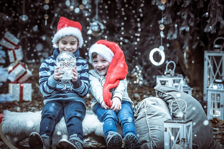 Auf diesem Bild sehen Sie eine Familie, die frohlich in weihnachtslicher Stimmung umgeben von Geschenken, Tannenbäumen und Kugeln mit Weihnachtsmützen in die Kamera lächelt