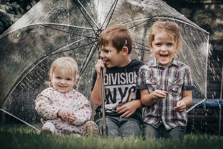 Auf diesem Bild sehen Sie drei Geschwister, die auf einer Wiese unter einem transparenten Regenschirm sitzen, als es regnet