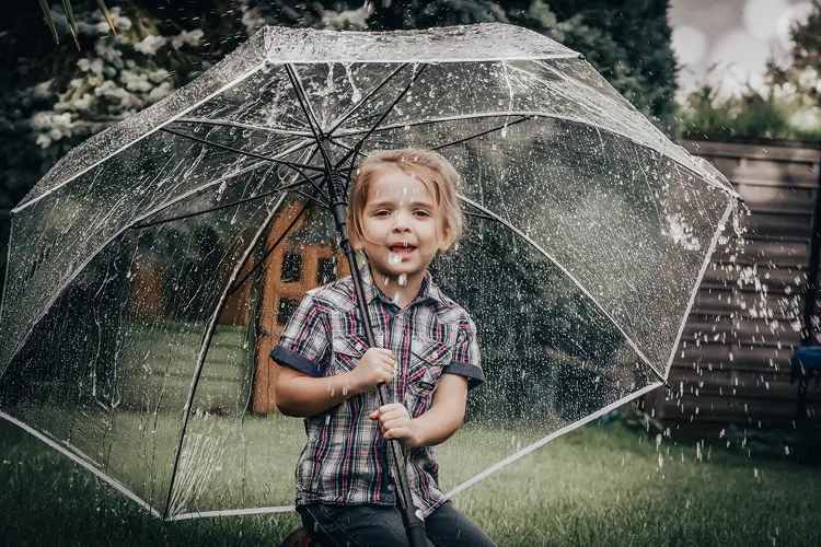 Auf diesem Bild sehen Sie ein kleines Mädchen, was auf einer Wiese unter einem transparenten Regenschirm sitzt, als es regnet